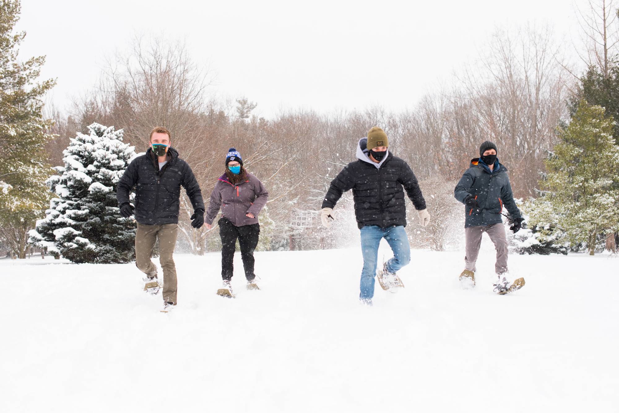 Image of 4 students wearing masks in the snowy outdoors with snow shoes on walking towards the foreground.